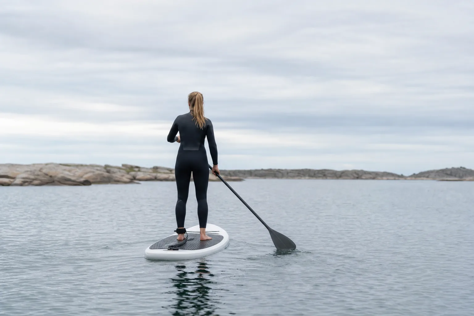 Person i våtdräkt sedd bakifrån stående på en SUP-bräda på lugnt vatten